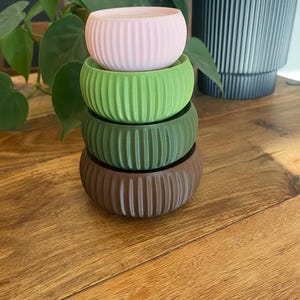 May include: Four stacked, ribbed, round planters in shades of pink, green, and brown. The planters are on a wooden surface, with green plants in the background. The planters are a decorative item.