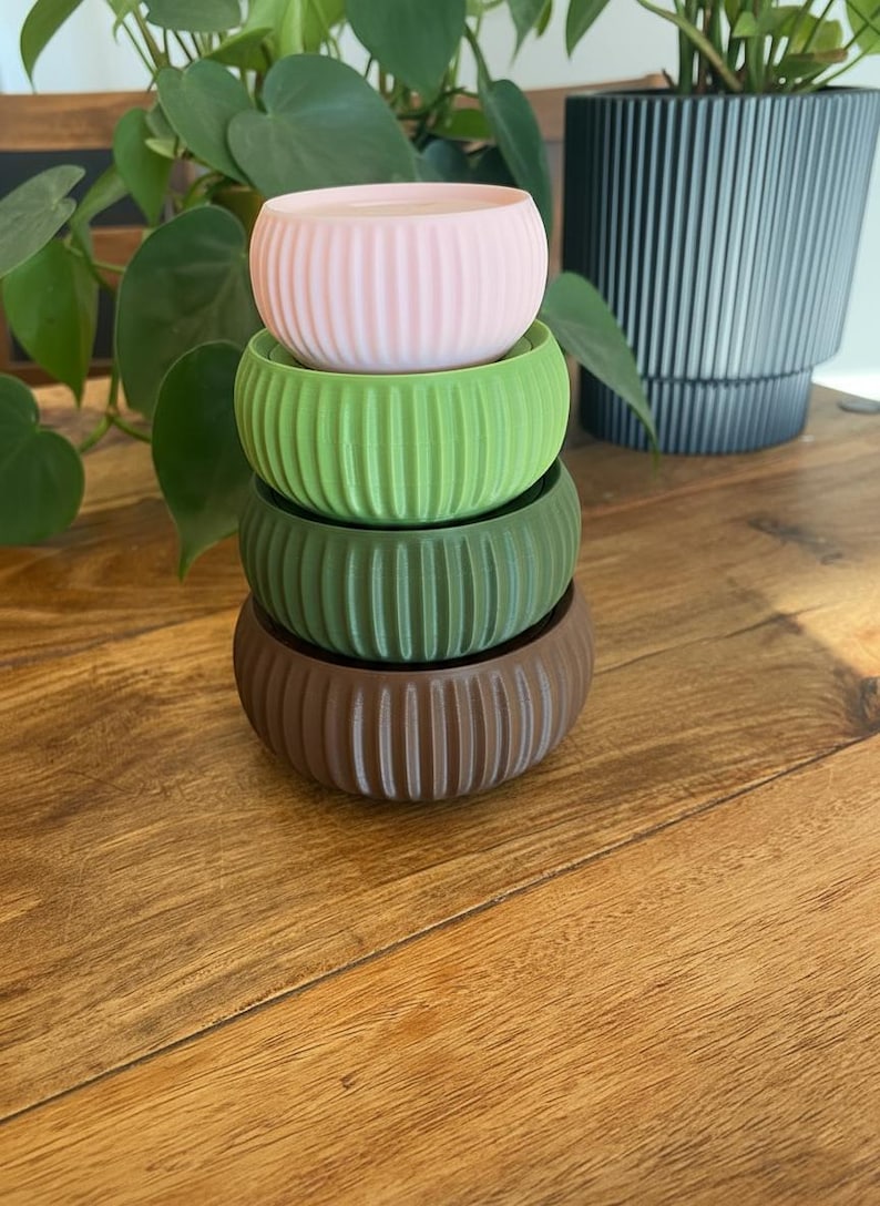 May include: Four stacked, ribbed, round planters in shades of pink, green, and brown. The planters are on a wooden surface, with green plants in the background. The planters are a decorative item.