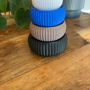 May include: A stack of four small, ribbed planters in white, blue, tan, and black. The planters are cylindrical with vertical grooves and sit on a wooden surface. A green plant is visible in the background.