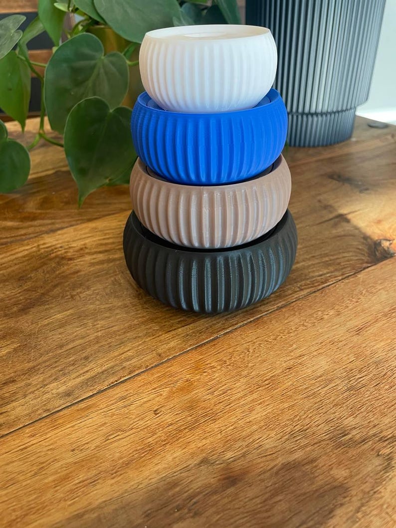 May include: A stack of four small, ribbed planters in white, blue, tan, and black. The planters are cylindrical with vertical grooves and sit on a wooden surface. A green plant is visible in the background.