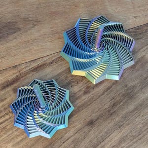 Könnte beinhalten: Zwei schillernde, sternförmige Dekorationsgegenstände mit einem Spiraldesign. Jeder Gegenstand hat einen zentralen, vertikalen Pfosten. Die Objekte sind in Blau-, Grün- und Lilatönen gehalten. Sie befinden sich auf einer Holzoberfläche.