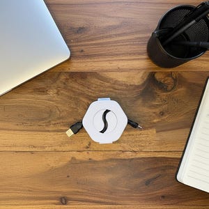 Könnte beinhalten: Ein weiß-schwarzer sechseckiger USB-Hub mit ausziehbarem Kabel befindet sich auf einem Holzschreibtisch. Ein Laptop, ein Stifthalter mit Stiften und Bleistiften sowie ein Notizbuch mit einem Stift befinden sich ebenfalls auf dem Schreibtisch.