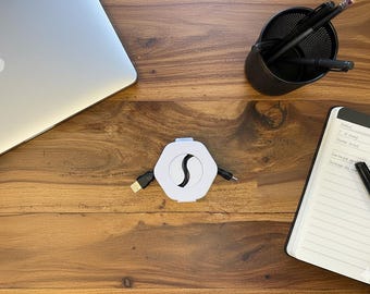 Może przedstawiać: Biało-czarny sześciokątny koncentrator USB z chowanym kablem znajduje się na drewnianym biurku. Na biurku znajduje się również laptop, pojemnik na długopisy z długopisami i ołówkami oraz notatnik z długopisem.