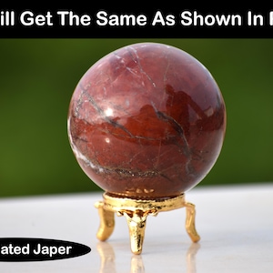 Può includere: Una sfera lucida di diaspro brecciato, principalmente rossa con venature scure, esposta su un supporto dorato ornato a tre gambe. Il testo "You Will Get The Same As Shown In Photos" è in alto e "Brecciated Japer" in basso.