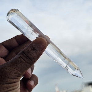 Puede incluir: Una varita de cristal de cuarzo transparente con una parte superior redondeada y un extremo puntiagudo. El cristal se sostiene en una mano sobre un fondo de cielo nublado. El texto "AGATESTONE" es visible en el cristal.