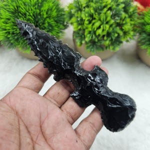 May include: A black obsidian arrowhead with a textured surface. The arrowhead is held in a hand and has a sharp, pointed tip. The arrowhead is a dark, glossy black color.