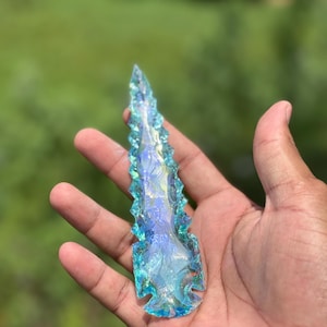 May include: A hand holds a translucent, iridescent arrowhead. The arrowhead is a vibrant mix of blues and greens, with a jagged edge and a pointed tip. The background is a blurred green.