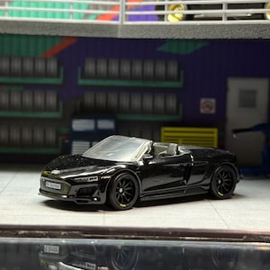 Puede incluir: Un coche de juguete descapotable negro con ruedas negras y un parabrisas plateado. El coche es un modelo en miniatura, probablemente de colección. El fondo sugiere un garaje o taller, con otros coches de juguete visibles.