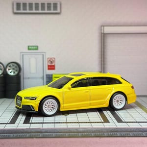 May include: A vibrant yellow model car with white wheels is positioned in a garage setting. The car features a streamlined design and a panoramic sunroof. The background includes a garage door, tyres, and overhead lighting.