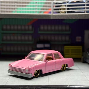 May include: A pink die-cast toy sedan featuring a silver front bumper, round headlights, and gold-colored five-spoke wheels. The vehicle has a boxy body style with a tan interior visible through the clear windows, presented in a garage setting.