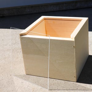May include: A light-colored wooden box with a clear acrylic panel. The box is square with an open top, revealing the natural wood grain inside. The acrylic panel is positioned to the left of the box.
