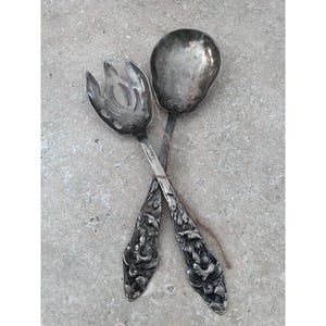 May include: A silver serving spoon and fork set, crossed over each other. The spoon has a rounded bowl, and the fork has three tines. Both utensils have ornate, detailed handles with a floral design.