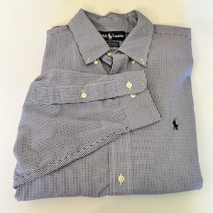 Könnte beinhalten: Ein gefaltetes Hemd mit blau-weißem Vichy-Karo-Muster. Es hat einen Button-down-Kragen und geknöpfte Manschetten. Ein kleines, dunkelblaues Logo ist unten links aufgestickt. Das Etikett lautet "Ralph Lauren".