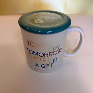 May include: White ceramic mug with a teal rim and lid. The lid features a floral design and text. The mug has a curved handle and text in various colors: "Yesterday is history, tomorrow is mystery, today is a gift."