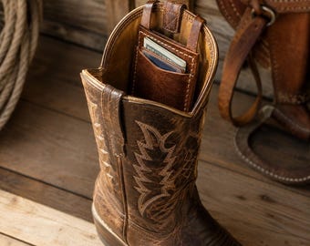 May include: A brown leather cowboy boot featuring decorative cream-coloured stitching on the shaft. A small, matching brown leather pocket is attached to the interior upper rim of the boot, containing a stack of paper currency and a card.