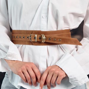 May include: A wide tan leather belt featuring a series of metal grommets along the length, a rectangular buckle, and two circular metal rings. The belt is worn over a crisp white button-down shirt with long sleeves and a visible center button.
