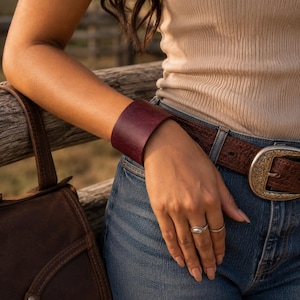 Puede incluir: Una pulsera de brazalete ancha de cuero color burdeos profundo, lisa y mate. La pulsera presenta un diseño sólido y minimalista que rodea completamente la muñeca, sin cierres, hebillas ni herrajes visibles en su superficie.