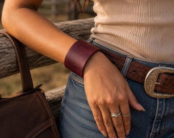 May include: A wide, smooth, deep burgundy leather cuff bracelet worn on the wrist. The bracelet features a solid, minimalist design with a matte finish, encircling the arm completely without any visible clasps, buckles, or additional hardware.