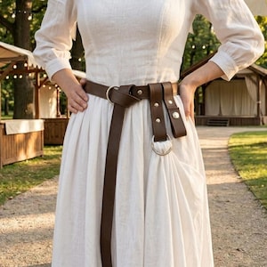 May include: A long-sleeved white dress featuring a fitted bodice and a full skirt, cinched at the waist with a dark brown leather belt. The belt includes a silver-toned metal ring attachment, multiple rivets, and a long strap that hangs down the front.