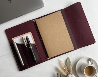 May include: A deep burgundy leather notebook cover featuring an open design with a tan paper notebook insert, two pen slots containing a silver pen and a black pen, and a small rectangular notepad tucked into a side pocket on the left interior.