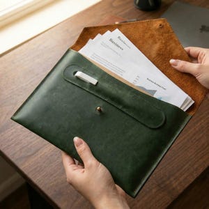 以下が含まれることがあります： フラップ開閉式で、金属製のスタッドボタンと白いペンホルダーが付いたダークグリーンのレザー製ドキュメントフォルダー。内側はナチュラルなタンカラーのスエード張りで、Businessという文字が印刷された白い紙が数枚入っています。