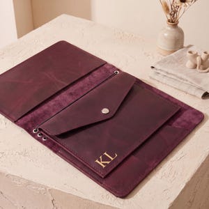 May include: A deep burgundy leather portfolio featuring an envelope-style pocket with a silver snap closure and the gold embossed initials KL. The interior includes multiple slots and metal eyelets for binding, showcasing a smooth, matte finish.
