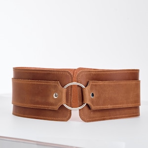 May include: A wide brown leather belt featuring a smooth matte finish and a central silver-toned metal ring closure. The belt is constructed with two main panels joined by the ring, secured with silver-toned rivets on each side of the hardware.