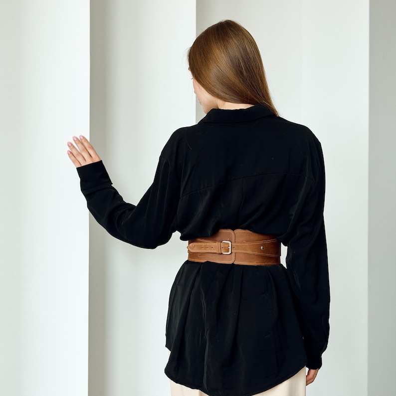 May include: A wide tan suede corset-style belt featuring a central silver-toned metal buckle and multiple adjustment holes, worn over a long-sleeved black button-down shirt with a collared neckline and a loose, relaxed fit.