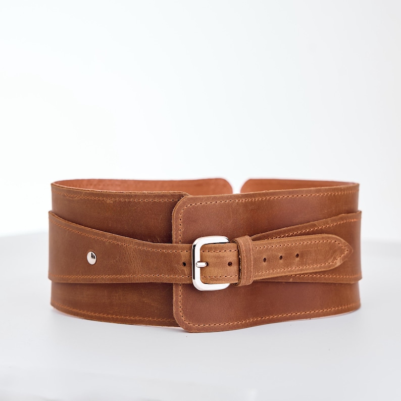 May include: A wide brown leather belt featuring a layered design with a silver-toned metal buckle. The belt has visible stitching along the edges and a single silver metal rivet on the left side, with multiple adjustment holes on the strap.