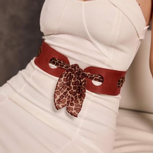 Pode incluir: Um cinto de couro vermelho com um lenço com estampa de leopardo amarrado em um laço. O cinto tem um design largo com recortes decorativos e é usado sobre um vestido branco. A estampa de leopardo adiciona um contraste elegante.