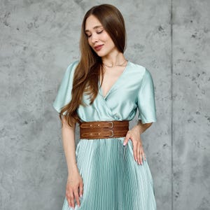 May include: A light blue dress with a pleated skirt and a wrap-style bodice. A wide brown leather belt with two silver buckles cinches the waist. The dress has short sleeves and a V-neckline.