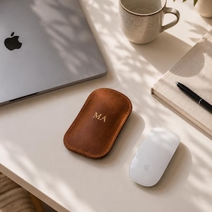 Op de afbeelding: Een bruin lederen etui met afgeronde hoeken en zichtbare stiksels, voorzien van de goudkleurige letters MA op de voorzijde. Het etui ligt op een plat oppervlak naast een zilveren laptop, een witte computermuis, een mok en een notitieboek.