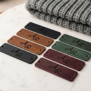 May include: A collection of rectangular leather tags in various colors, including black, brown, green, and burgundy. Each tag is embossed with the initials "AR" in a cursive font. A folded gray knitted item is in the background.