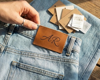 May include: Light blue denim jeans featuring a rectangular tan leather patch on the back waistband with the letters AR embossed in a cursive script. A stack of several small, square, beige and white paper tags is attached to the jeans.