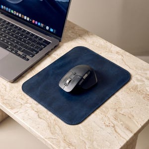 Op de afbeelding: Een rechthoekige donkerblauwe leren muismat met afgeronde hoeken en gestikte randen. Een zwarte ergonomische draadloze computermuis met een metalen scrollwiel en zijknoppen is in het midden van de muismat geplaatst.