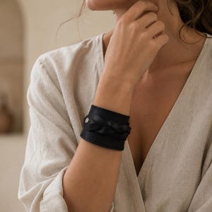 May include: A wide black leather cuff bracelet featuring a central braided leather detail and a single silver-toned metal stud closure. The bracelet has visible stitching along the top and bottom edges, creating a structured and solid band design.