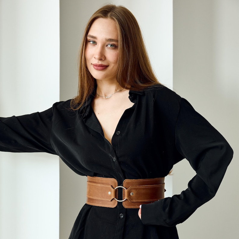 May include: A wide brown leather belt featuring a central silver-toned metal ring buckle. The belt has a structured design with visible stitching along the edges and two silver rivets securing the leather to the circular metal hardware.
