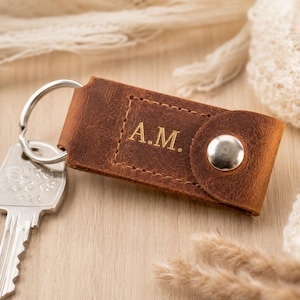 May include: A brown leather keyring featuring a silver metal snap button and a silver split ring attached to a metal key. The leather surface is embossed with the letters A.M. and has visible stitching along the rectangular centre panel.