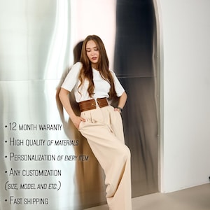May include: A white short-sleeved t-shirt paired with high-waisted beige trousers and a wide brown leather corset-style belt featuring a central metal ring buckle. The outfit is clean and neutral, highlighting the structured fit of the wide-leg trousers.