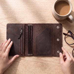 May include: A dark brown leather notebook cover featuring an interior zippered pocket, three vertical elastic bands for securing inserts, and a right-side pocket. The cover includes three metal snap fasteners along the edge for secure closure.