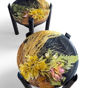 May include: Two small, round tables with black legs and tops. The table tops are black and feature dried flowers and grasses encased in a clear resin. The flowers are yellow, pink, and white, and the grasses are tan.