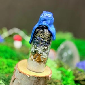 May include: A clear glass vial filled with layers of dried herbs, lavender, and white crystals, topped with a sculpted blue wax seal. The vial is positioned upright on a small, cylindrical wooden log slice, showcasing a natural, rustic aesthetic.