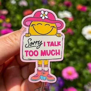 May include: A pink and yellow badge reel with a smiling cartoon character wearing a hat with a flower. The character holds a sign that reads "Sorry I talk too much". The badge reel has a silver clip.