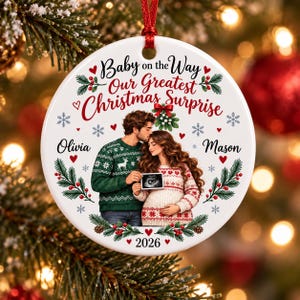 May include: A round white ceramic Christmas ornament featuring an illustration of a couple in holiday sweaters holding an ultrasound image. The text reads Baby on the Way Our Greatest Christmas Surprise, Olivia, Mason, and 2026, with holly and snowflakes.