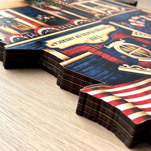 May include: A multi-layered wooden wall art piece featuring a patriotic theme with red, white, and blue colors. The design includes an American flag, architectural details, and a banner with the text THE UNITED STATES OF AMERICA printed.