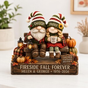 May include: A decorative figurine of two gnomes sitting on a bench, wearing striped hats labeled George and Helen. The gnome on the left holds a lantern, while the other holds a mug labeled WARM & COZY. The base reads FIRESIDE FALL FOREVER.