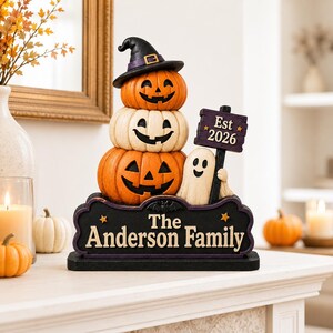 May include: A decorative Halloween tabletop sign featuring a stack of three pumpkins, the top one wearing a black witch hat. A small ghost holds a purple sign reading Est 2026. The base is a black plaque with the text The Anderson Family in white.
