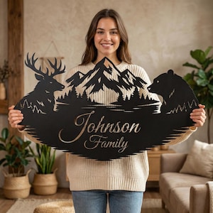 Może przedstawiać: Czarny metalowy znak rodzinny z sylwetką jelenia, gór i niedźwiedzia. Znak zawiera nazwisko rodziny "Johnson Family" w ozdobnej czcionce. Znak ma motyw natury.