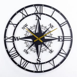 Op de afbeelding: Een grote ronde wandklok met een zwart metalen frame en Romeinse cijfers langs de buitenrand. Het midden toont een zwart kompasroosontwerp met de windstreken N, NE, E, SE, S, SW, W en NW, en voorzien van goudkleurige wijzers.