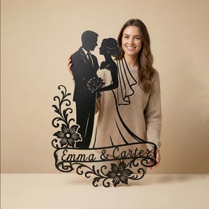 Puede incluir: Letrero de boda de metal negro con una silueta de novios, con el texto "Emma & Carter" en una pancarta. El letrero tiene detalles florales y de volutas, ideal para la decoración de bodas.